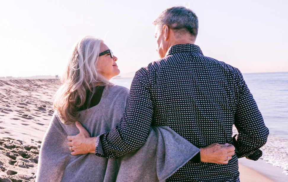 an elderly couple walks on beach and looks at each other