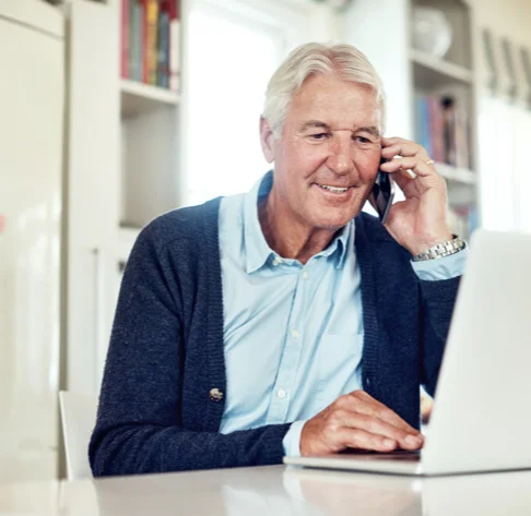 an elderly man sits and uses laptop while talking on mobile phone